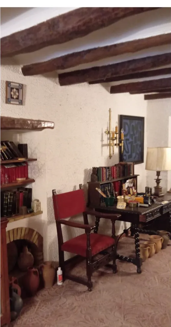 Estancia con vigas de madera y suelo de barro cocido antes del vaciado: escritorio, sillón, estanterías con libros y cántaros de cerámica junto a la chimenea