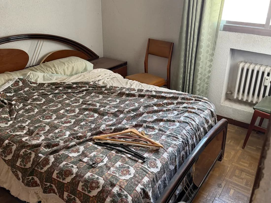 Dormitorio antes del vaciado: cama de matrimonio con cabecero de madera y colcha estampada, mesilla, una silla y perchas sobre la cama, con el radiador en su hueco junto a la ventana