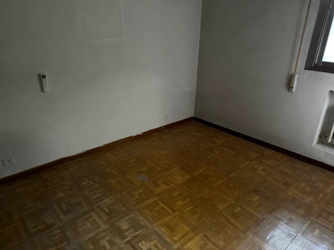 El mismo dormitorio ya vacío tras el vaciado, con el suelo de parqué despejado y el hueco del radiador junto a la ventana