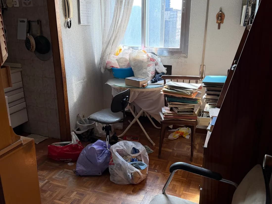 Habitación de un piso antes del vaciado: bolsas en el suelo, una mesa camilla con ropa y cajas, montones de carpetas y papeles sobre una silla, y una silla de oficina junto a la puerta de la cocina