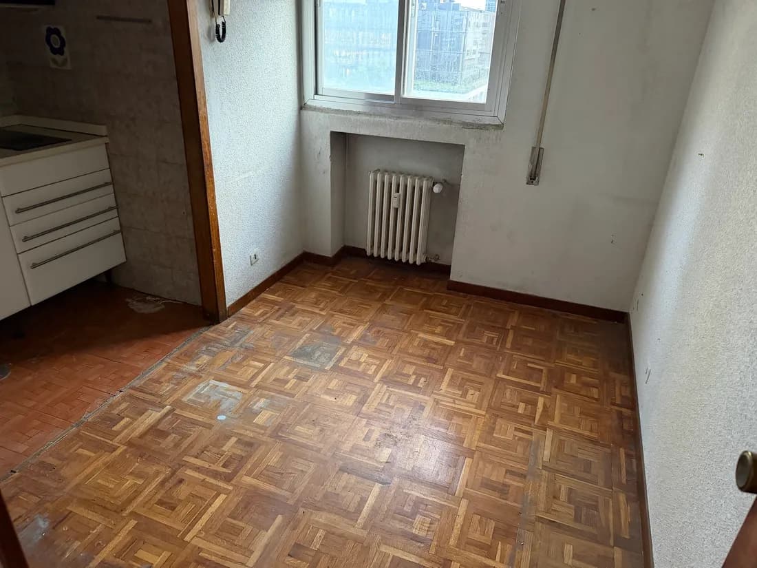 La misma habitación ya vacía tras el vaciado, con el suelo de parqué barrido, el radiador en su hueco bajo la ventana y la puerta de la cocina al fondo